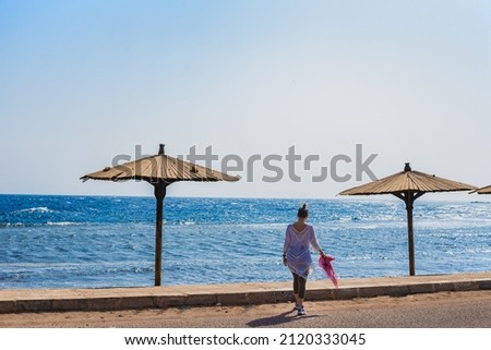 Exotic country vacation island beach. Young woman in white sun dress walking along sea coast. Horizontal sea shore with female tourist .copy space. Clear blue sky over ocean waves.