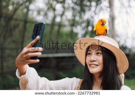 Exotic pet, Happy traveler asian woman with mobile phone selfie with Sun conure parrot bird on her head at day in Thailand Zoo, Sun conure parrot is famous bird pet in Thailand