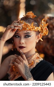 Exotic Woman In Her Bold Makeup While Wearing Golden Accessories On Her Head And Neck While In A Black Dress Inside The Forest