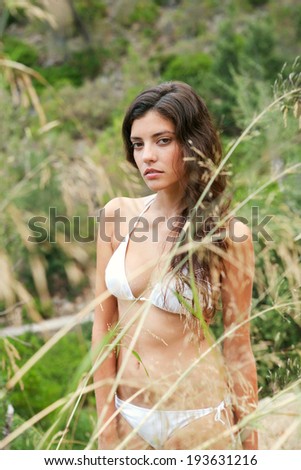 Exotic young woman standing in the vegetation of a beach wearing a bikini and being thoughtful while relaxing on a summer vacation with a perfect body, outdoors. Travel and beauty lifestyle.