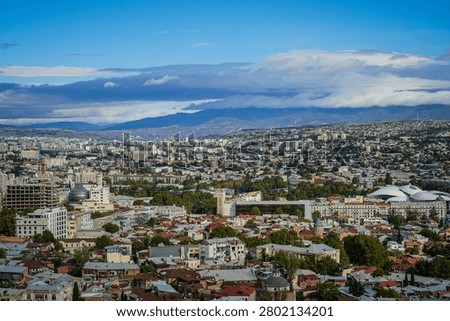 An expansive elevated view captures the vibrant city of Tbilisi, Georgia, with its diverse architecture and lush greenery spreading towards distant mountains. The modern Rike Park Concert Hall and Exhibition Center stands out among the urban landscape under a partly cloudy blue sky.