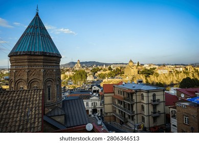 An expansive high-angle view captures the architectural beauty of Tbilisi, Georgia, with the brick Sioni Cathedral Bell Tower dominating the foreground. In the mid-ground, the Metekhi Church stands atop a cliff overlooking the Kura River, while the distant Holy Trinity Cathedral of Tbilisi crowns a hill under a clear blue sky.