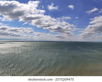 Expansive ocean view under a vibrant blue sky with scattered clouds, sunlight reflecting on rippling waves. A serene and vast seascape showcasing nature's beauty.