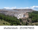 Expansive open pit gold mining operation in Colorado with heavy machinery working on a sunny day amidst lush greenery and distant mountains