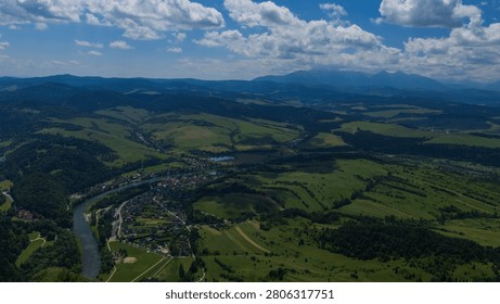 Expansive panoramic landscape featuring a winding river flowing through a lush green valley with farmland, forests, and scattered rural settlements. Distant mountain ranges rise beneath a vivid blue sky filled with dramatic cumulus clouds, creating a breathtaking summer nature scene viewed from above