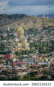 An expansive panoramic view of the Holy Trinity Cathedral of Tbilisi, also known as Sameba Cathedral, standing prominently with its iconic golden dome amidst the bustling cityscape of Tbilisi, Georgia. The surrounding urban landscape features numerous houses and green trees, with rolling hills under a partly cloudy sky in the background.