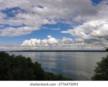 Expansive river landscape under a dramatic sky filled with white clouds, with calm water reflecting the blue sky and green forested shoreline, capturing the serene beauty of summer nature