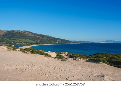 Expansive sand dunes lead to the deep blue sea in Tarifa, Spain. Rolling hills with wind turbines are visible, with the African coastline in the distance. - Powered by Shutterstock