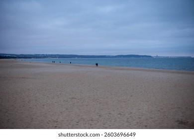 Expansive sandy beach with distant figures strolling, calm sea, and a hazy skyline create a tranquil, overcast coastal scene under a soft gray sky
