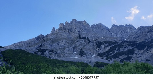 Expansive view of a formidable mountain range under a vast blue sky, featuring jagged rocky peaks, sparse vegetation, and remnants of snow. Evokes the powerful beauty of alpine terrain.