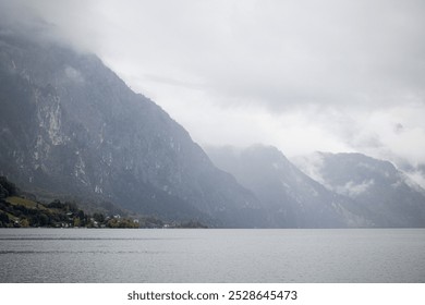 Expansive view of a misty mountain range by a calm lake under an overcast sky. The mountains rise dramatically, creating a moody and tranquil atmosphere - Powered by Shutterstock