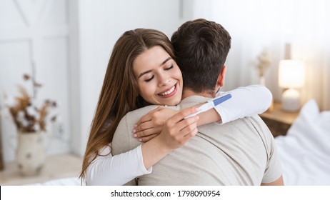 Expect a baby. Happy young woman hugging husband and looking at positive pregnancy test in interior of bedroom, panorama, free space