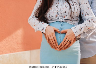 Expectant couple making heart shapes with hands while standing against pink wall - Powered by Shutterstock