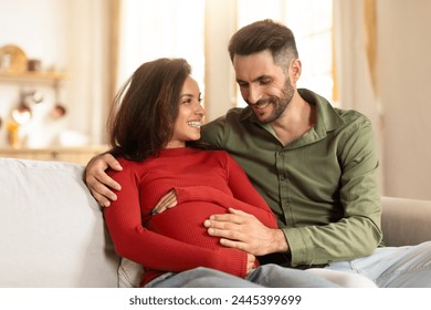 An expectant couple shares a tender moment on the sofa, with the focus on the pregnant woman's belly - Powered by Shutterstock