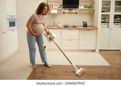 The expectant female is smiling while using a handheld vacuum to clean the kitchen. The pregnant woman is happily doing housework, vacuuming the kitchen floor with a cleaner