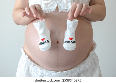 Expectant mother holds a pair of cute newborn baby socks with love heart mama design over her beautiful pregnancy belly baby bump. Pregnancy, motherhood, expectation concept. - Powered by Shutterstock