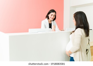 Expectant mother touching her belly while talking with a female receptionist. Female worker checking in a patient at the gynecologist office