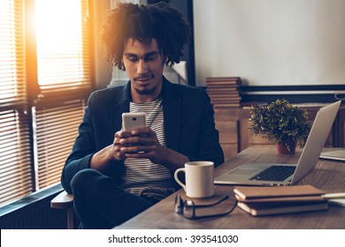 Expected message. Young handsome African man using his smartphone with smile while sitting at his working place