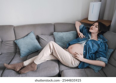 Expecting mother lying on gray sofa, smiling, wearing comfortable clothes. - Powered by Shutterstock