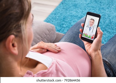 Expecting Pregnant Woman Sitting On Sofa Video Conferencing With Doctor On Mobile Phone