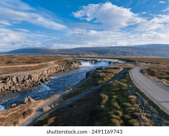Experience the breathtaking beauty of Iceland from above, showcasing a light blue waterfall, rocky terrain, and a winding road under a bright blue sky. - Powered by Shutterstock