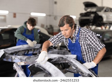 Experienced auto body technician in blue overalls focused on smoothing surface of car parts using sanding block in vehicle repair workshop, preparing for painting - Powered by Shutterstock