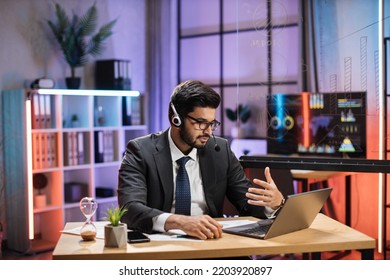 Experienced Bearded Indian Businessman, Economist In Suit Wearing Headset Sitting At Table With Laptop, Having Video Meeting At Evening Time At Office.