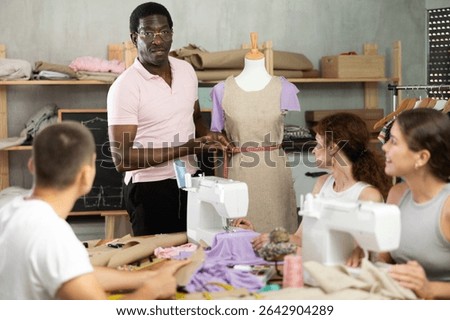 Experienced black sewing instructor explaining clothing construction techniques to group of engaged adult beginners working on garments during hands-on workshop in studio