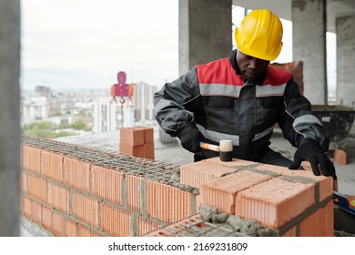 Experienced builder in workwear flattening surface of brick layer with special hammer while standing inside unfinished construction