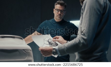 Experienced car designers and developers discuss the design of the car while standing infront of the sculpture of prototype car. Using the pencil sketches in a high tech car manufacturing company.