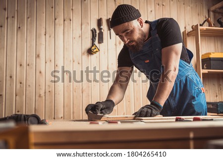 Experienced carpenter in work clothes and small business owner working in woodwork workshop,  using sandpaper for polishing  wooden bar  at worktable in workshop  