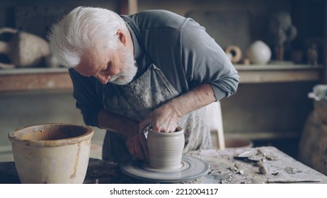 Experienced ceramist grey-haired bearded man is smoothing molded ceramic pot with wet sponge. Spinning throwing wheel, muddy work table and handmade clayware are visible. - Powered by Shutterstock