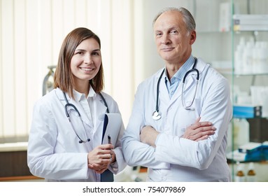 Experienced Clinician And His Young Assistant In Whitecoats Looking At Camera