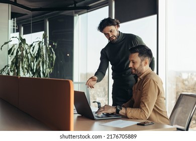 Experienced Colleague Explaining To Young Coworker Business Project Strategy At Contemporary Office, Copy Space. Two Entrepreneurs Discussing New Ideas And Strategies, Using Laptop Pc