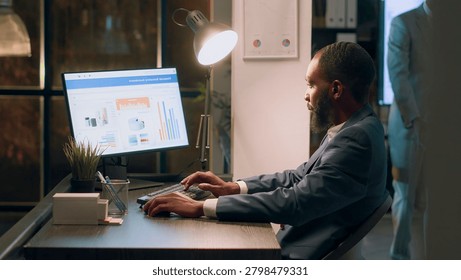 Experienced computer operator sitting at desk during nightshift, working on company accounting payroll reports and tables in modern office while coworker in background comes back from break
