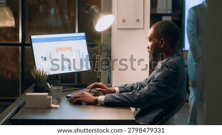 Experienced computer operator sitting at desk during nightshift, working on company accounting payroll reports and tables in modern office while coworker in background comes back from break