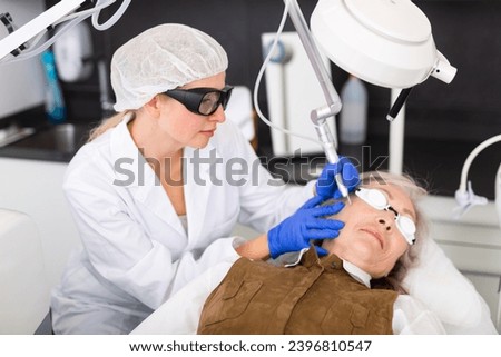 Experienced concentrated female cosmetologist performing laser facial treatment to elderly woman in aesthetic medicine office