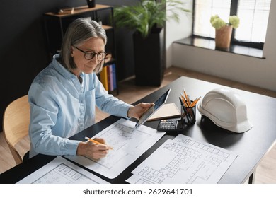 Experienced confident mature Caucasian female engineer architect using engineering tools and electronic devices while drawing blueprints sketch for real estate, sitting at her workplace office