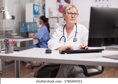 Experienced doctor using computer in clinic cabinet and nurse typing on pc in the background.
