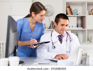 Experienced Doctor Working In Office With Female Assistant, Filling Up Medical Forms In Laptop