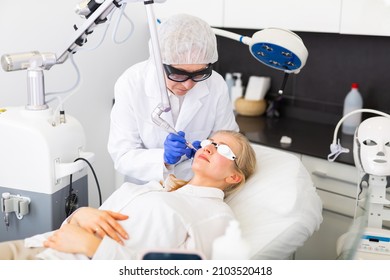 Experienced Elderly Female Cosmetologist Performing Laser Facial Treatment To Young Woman In Aesthetic Medicine Office