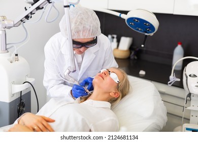 Experienced Elderly Female Cosmetologist Performing Laser Facial Treatment To Young Woman In Aesthetic Medicine Office