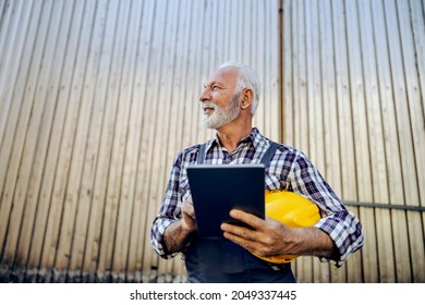 Experienced Engineer And Construction Manager At Work. A Older Man With Wears A Work Suit And Holds Documentation And A Protective Helmet In His Hands Project Monitoring And Control Of Job Realization