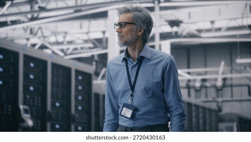 Experienced Engineer Overseeing Cloud System Integrity. Mature IT Specialist With Glasses Walks and Monitors Systems Clean, Bright Server Hall. Concept of Mobile System Management and Data Integrity.
