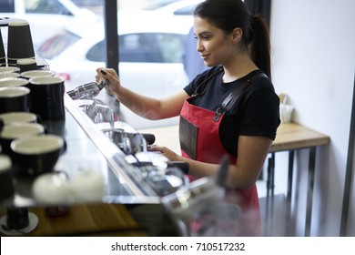 Experienced female barista enjoying process of coffee making satisfied with occupation in cafeteria.Pretty worker of cafe dressed in staff uniform preparing clients order on professional equipment
