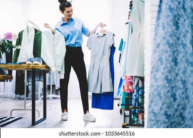 Experienced female dressmaker holding hangers with trendy apparel in hands and thinking on better variant of outfit working in fashion botique.Stylist of showroom choosing clothing for new image