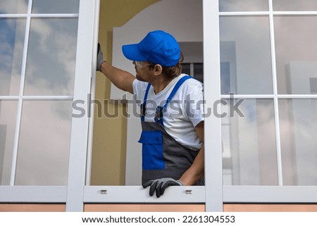 An experienced installer of plastic windows inspects the properly installed fittings. Man in work jumpsuit and a blue cap opened a window, checks the work done