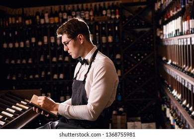 Experienced Male Wine Steward Doing Stocktaking Of Wine At Store, Writing In Notebook Some Notes In Dark Room With Rows Of Racks