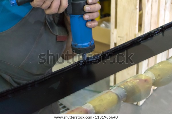 An experienced man in working clothes applies a\
black sealant to the glass removed from the car with a pneumatic\
pistol with a cartridge, pressing the trigger and moving along the\
edge in workshop