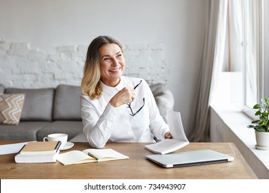 Experienced mature female chief editor holding glasses in one hand and book in other, laughing at sarcastic style of narration while reading novel of popular author. Writing and publishing concept